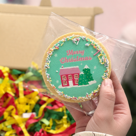 A hand holds a round decorated Christmas cookie in a clear plastic bag, with an open gift box filled with colorful paper shred in the background.